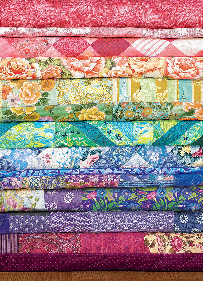 Cobble Hill 1000 Piece puzzle - Image of quilts stacked and folded in a rainbow of colors with different fabric patterns.